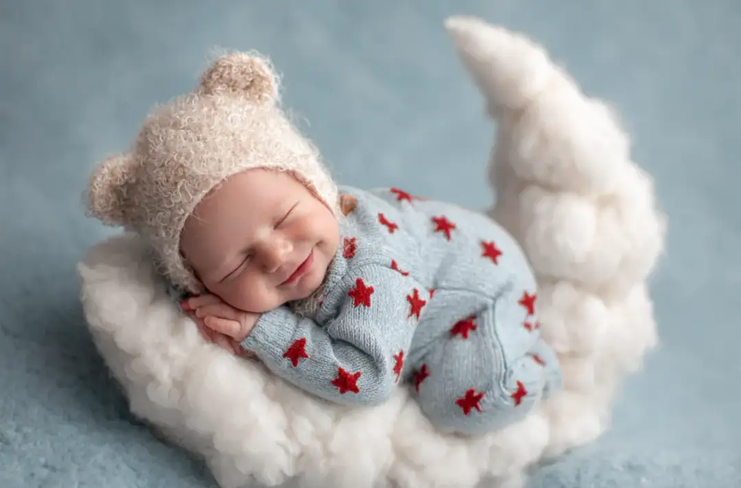 Uśmiechnięty noworodek w czapce z uszkami śpi na chmurce w kształcie księżyca.