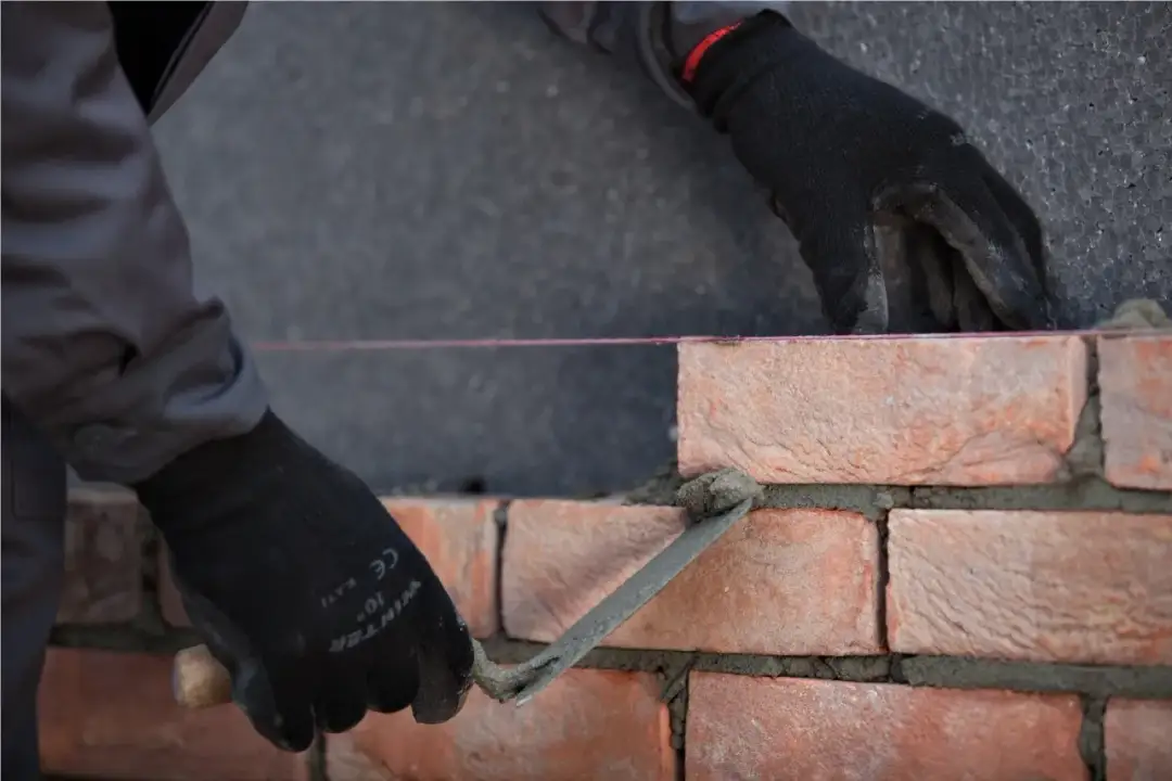 Ręce w czarnych rękawicach wykonują murowanie ścian działowych, układając cegły na zaprawie.