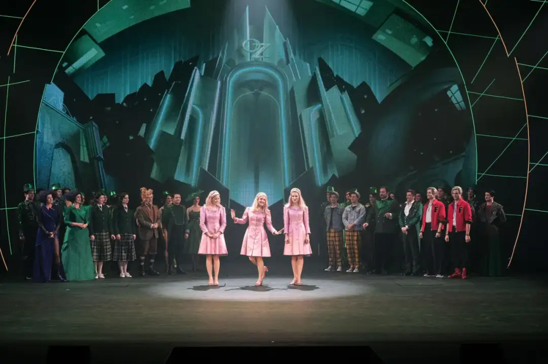 Trzy dziewczyny w różowych sukienkach na scenie, w tle zielony pałac Oz. To scena z musicalu "Wicked", który ma imponujący czas trwania.