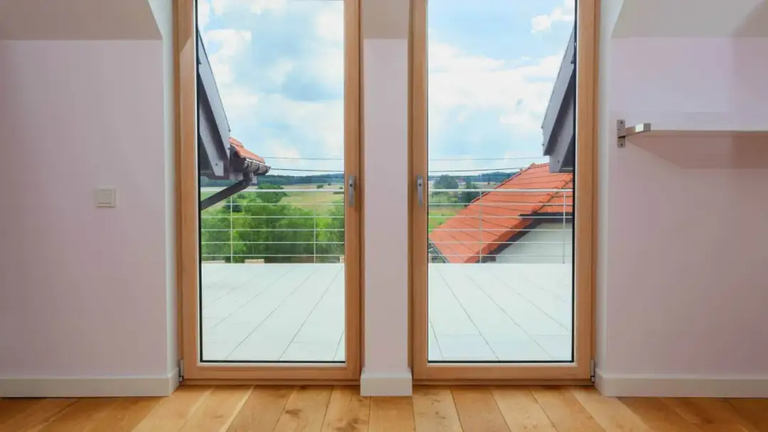 Drewniane okno balkonowe z widokiem na zielone pola i dachy. Zastanawiasz się, ile kosztuje okno balkonowe?