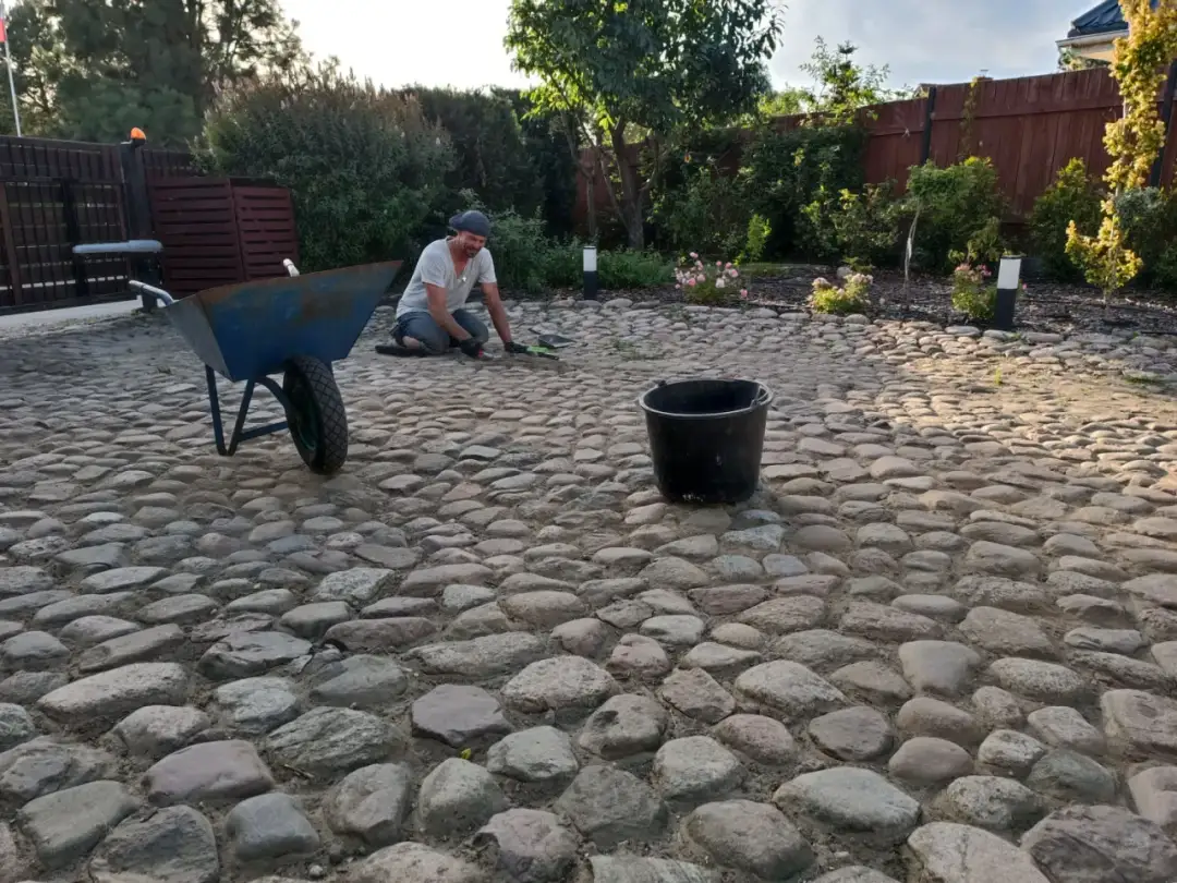 Mężczyzna pracuje przy budowie drogi z kamienia polnego. Obok stoi taczka i wiadro.