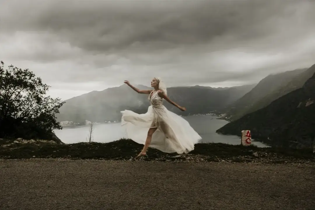 Panna młoda w zwiewnej sukni tańczy na tle gór i zatoki. Kiedy najlepiej kupić suknię ślubną?