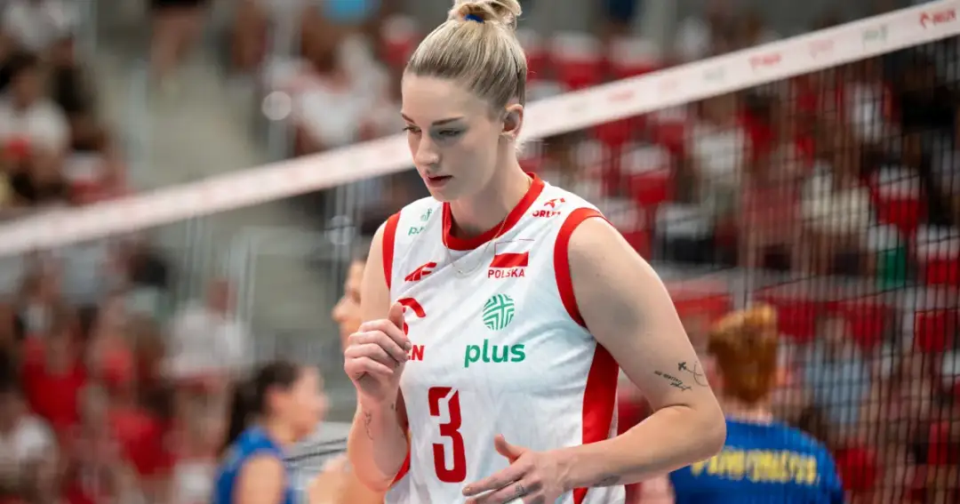 Siatkarka w białej koszulce z numerem 3, z orzełkiem i flagą Polski. Kibice na trybunach, atmosfera jak na meczu Ligi Mistrzyń. Tabela wyników w tle.