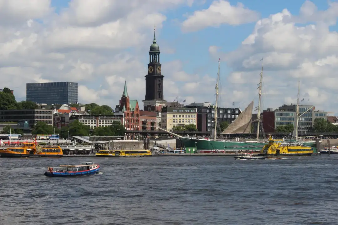 Widok na port w Hamburgu, gdzie statki i łodzie pływają po wodzie, a w tle widać miasto z wieżą kościelną. Czy Hamburg leży nad morzem? Tak, to miasto portowe!