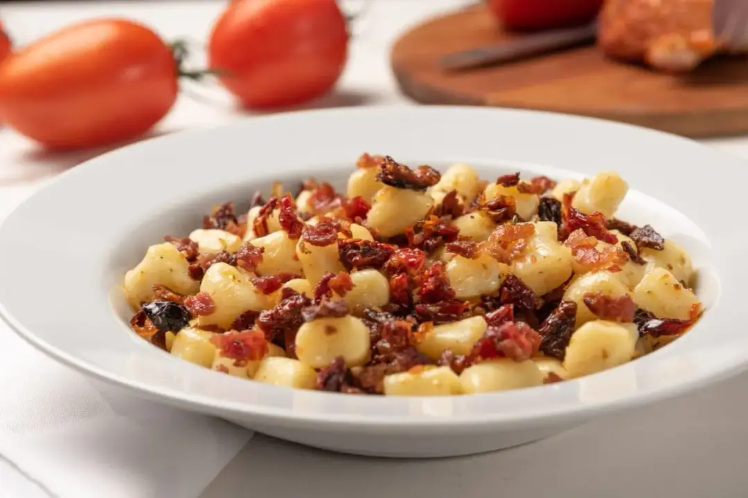 Pyszne gnocchi z boczkiem i suszonymi pomidorami na białym talerzu, w tle pomidory.