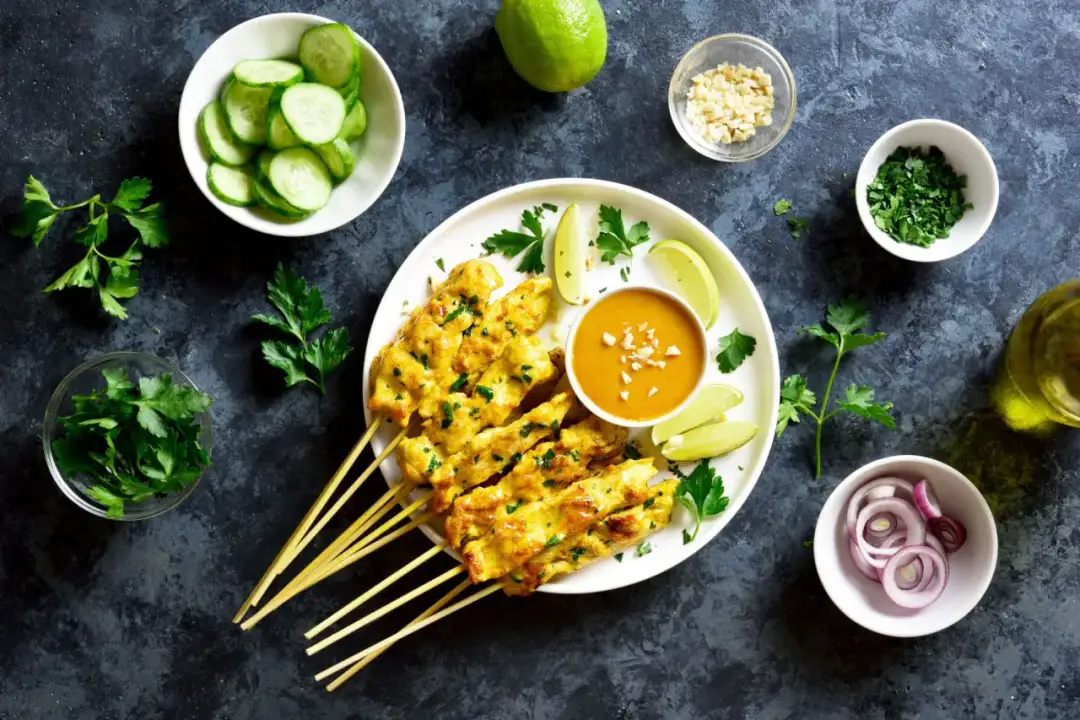 Pyszne satay z kurczaka na patyczkach, podane z sosem orzechowym, limonką, ogórkiem i posiekaną natką.