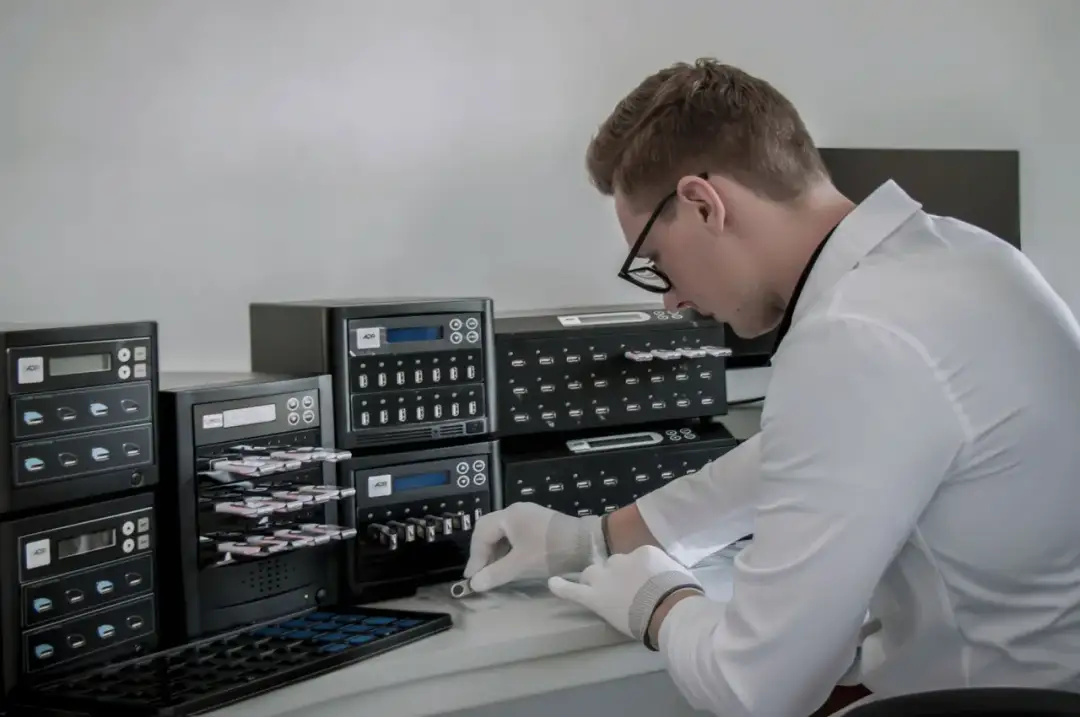 Mężczyzna w białej koszuli i rękawiczkach obsługuje kopiarkę pendrive'ów.