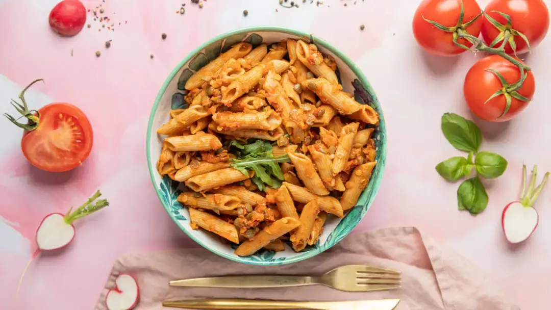 Miseczka pełna makaronu penne z sosem pomidorowym i soczewicą. Obok pomidory, rzodkiewki i bazylia. Idealna porcja, by ustalić, ile makaronu penne na osobę.