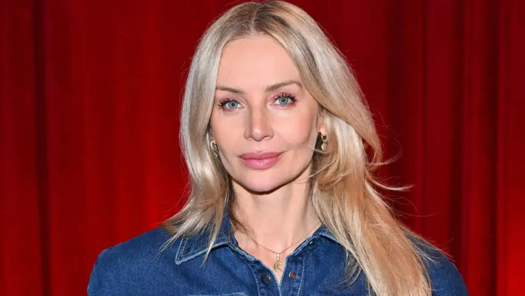 Agnieszka Woźniak-Starak w dżinsowej koszuli, z nowym partnerem u boku, prezentuje się promiennie na tle czerwonych zasłon.