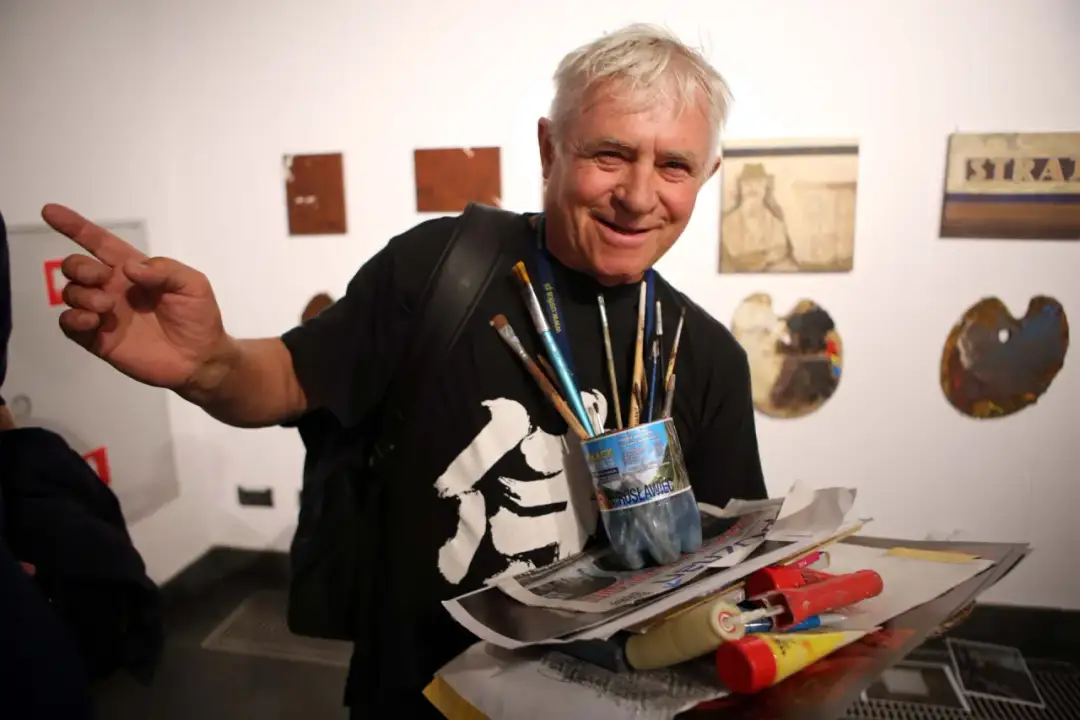 Uśmiechnięty Piotr C. Kowalski, artysta z pędzlami w butelce, prezentuje swoje prace.