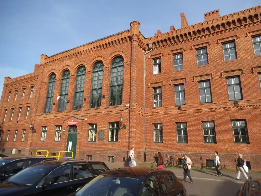 Ceglana kamienica z zielonymi oknami, gdzie w wieku 19 lat kończy się liceum. Ludzie idą chodnikiem.