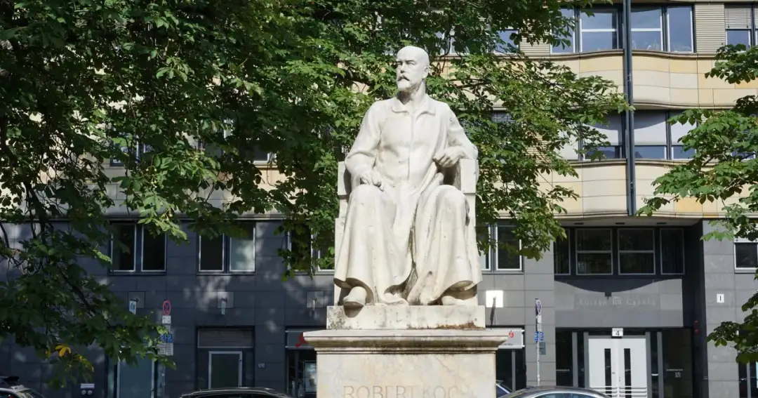Pomnik Roberta Kocha, który wynalazł szczepionkę na gruźlicę. Siedzi w długiej szacie, otoczony zielenią.