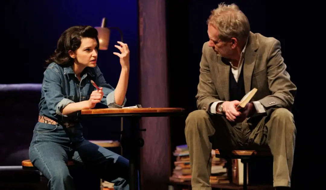 Kobieta w dżinsowej kurtce i mężczyzna w garniturze siedzą przy stoliku. To scena z przedstawienia, które może być prezentem urodzinowym.