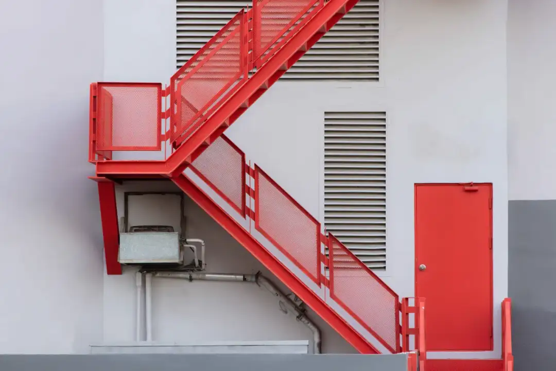Czerwone, metalowe schody zewnętrzne prowadzące na piętro, obok drzwi i kratki wentylacyjnej.
