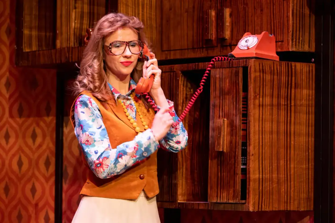 Kobieta w okularach i kwiecistej bluzce rozmawia przez czerwony telefon. Scena z spektaklu "Sylwester w Teatrze Warszawa".