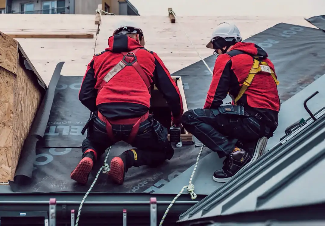 Dwóch dekarzy w czerwonych kurtkach i kaskach pracuje na dachu, zabezpieczonych linami.