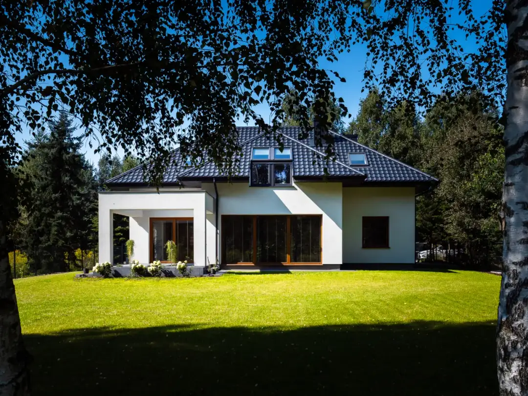 Nowoczesny, biały dom z dużymi oknami i zielonym trawnikiem, otoczony drzewami.