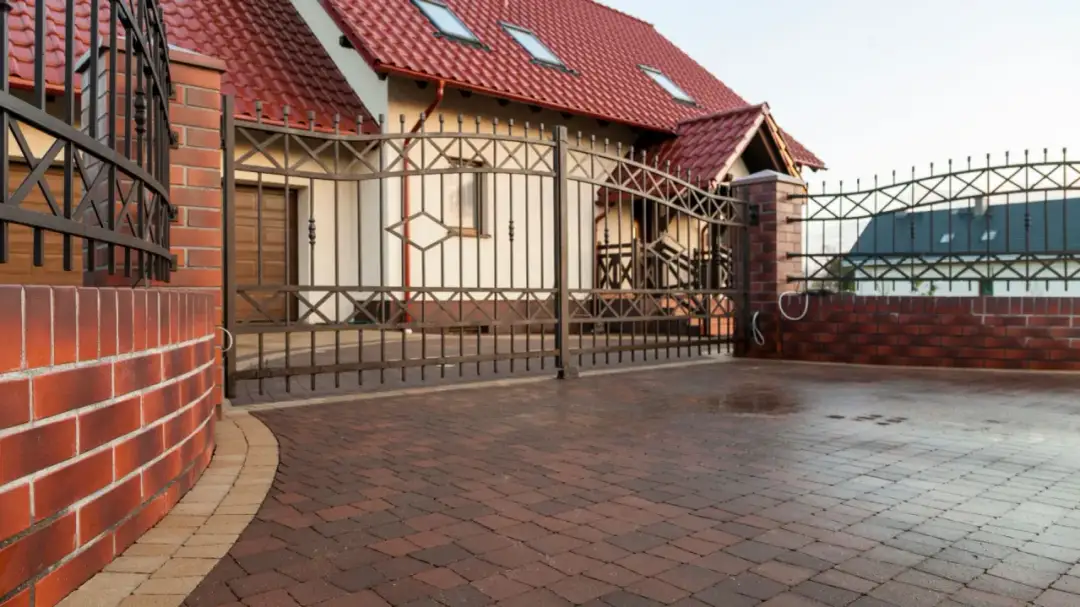 Długi wjazd na posesję z klinkierowej cegły i ozdobnej bramy. Dom z czerwonym dachem i oknami dachowymi.