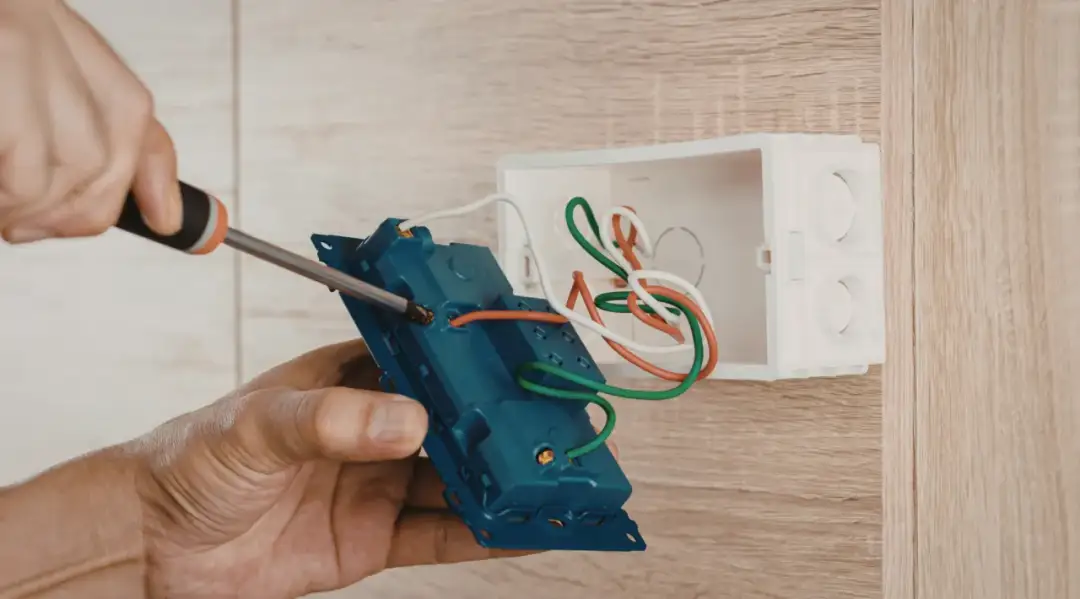 Dłoń przykręca śrubokrętem niebieski element instalacji elektrycznej z przewodami do białej puszki.