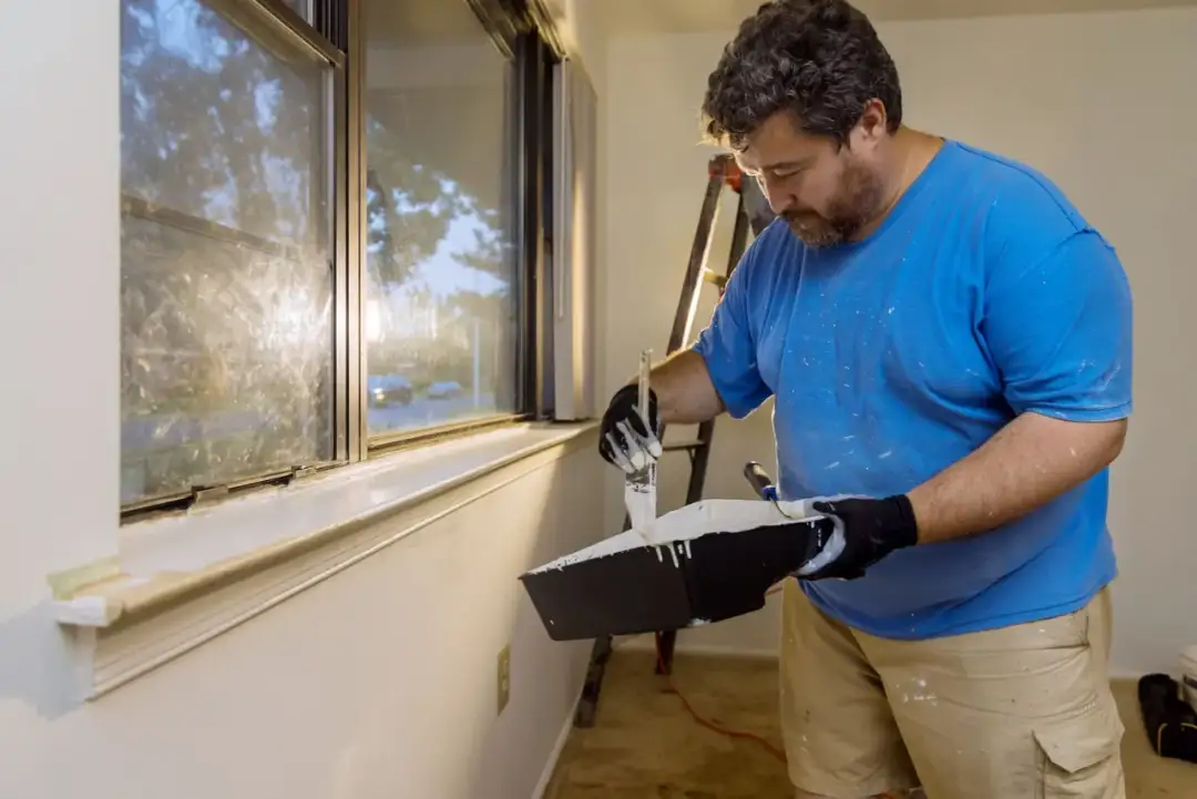 Mężczyzna w niebieskiej koszulce maluje pędzlem parapet na biało.