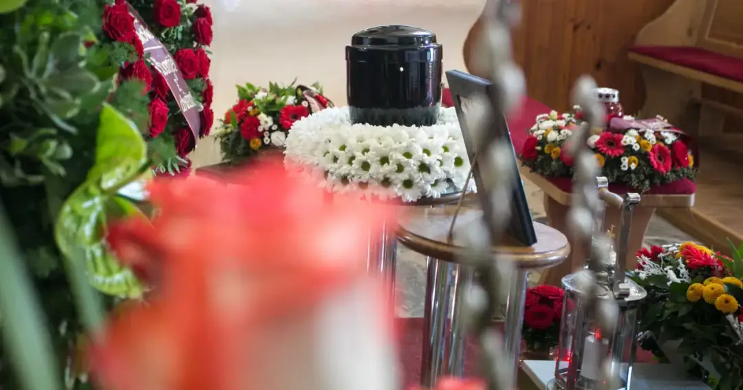 Czarna urna na wieńcu z białych kwiatów, otoczona bukietami. Czy kremacja boli zmarłego? Nie, to proces bezbolesny.