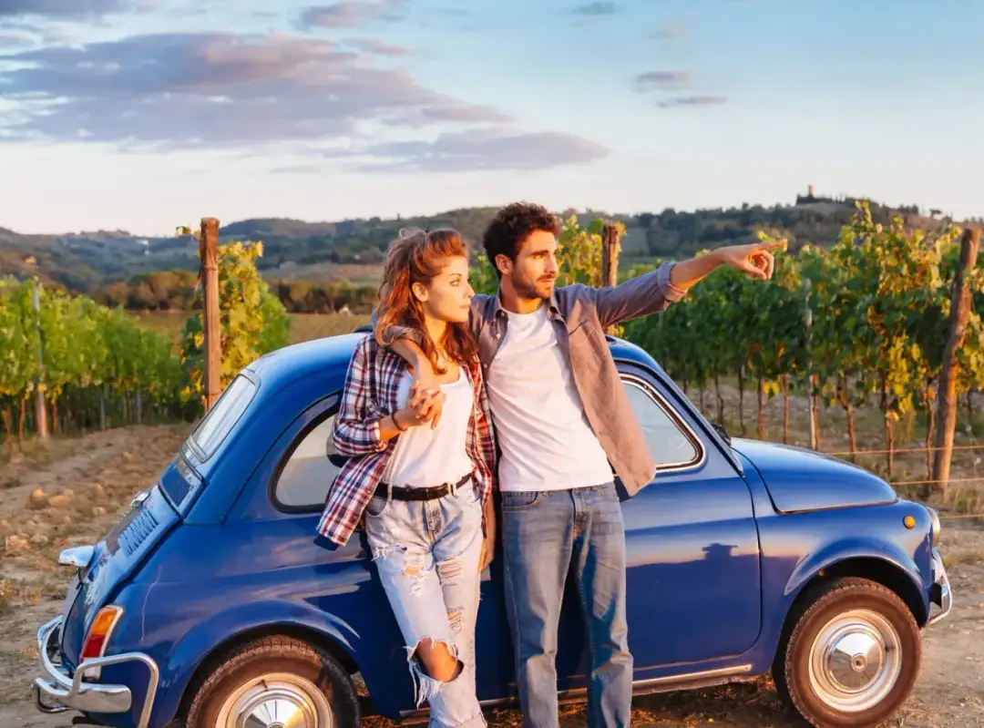 Para młoda przy niebieskim Fiat 500, planując podróż po Włoszech samochodem, wskazuje kierunek na tle winnic.