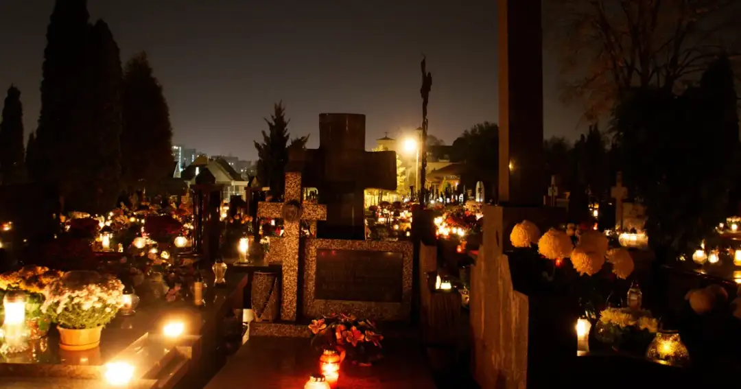 Cmentarz nocą, udekorowany zniczami i kwiatami, z widocznymi krzyżami i nagrobkami.