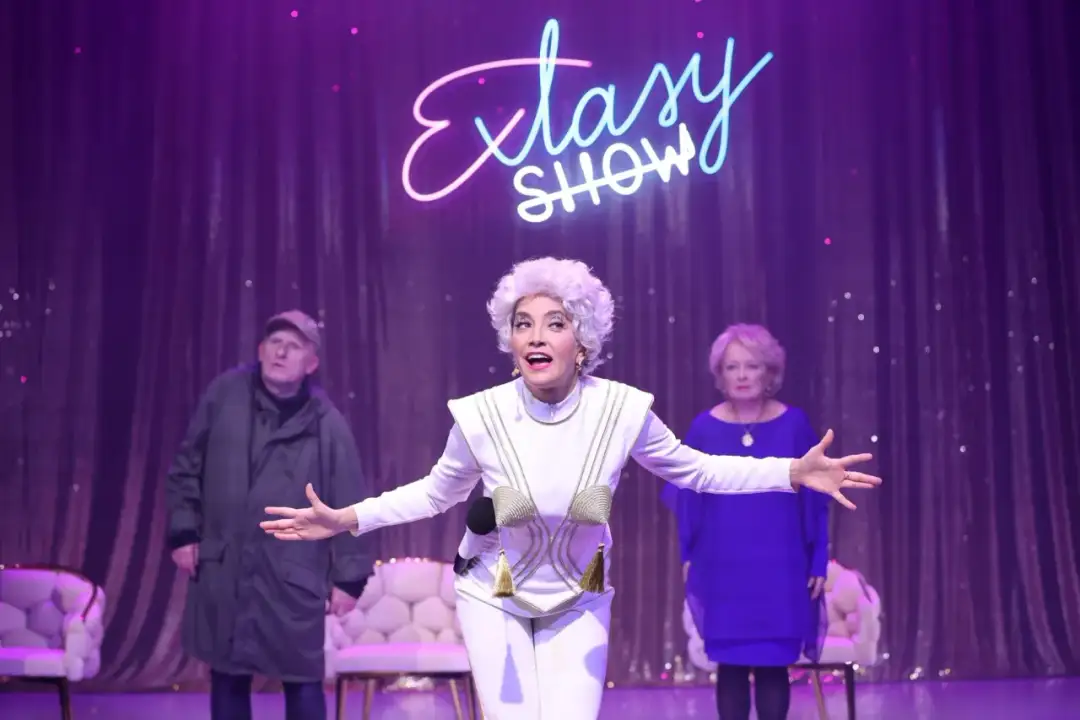 Voucher do teatru dla dwojga Warszawa: aktorka w białym stroju z frędzlami, mężczyzna w zielonej kurtce i kobieta w niebieskiej sukience na scenie.