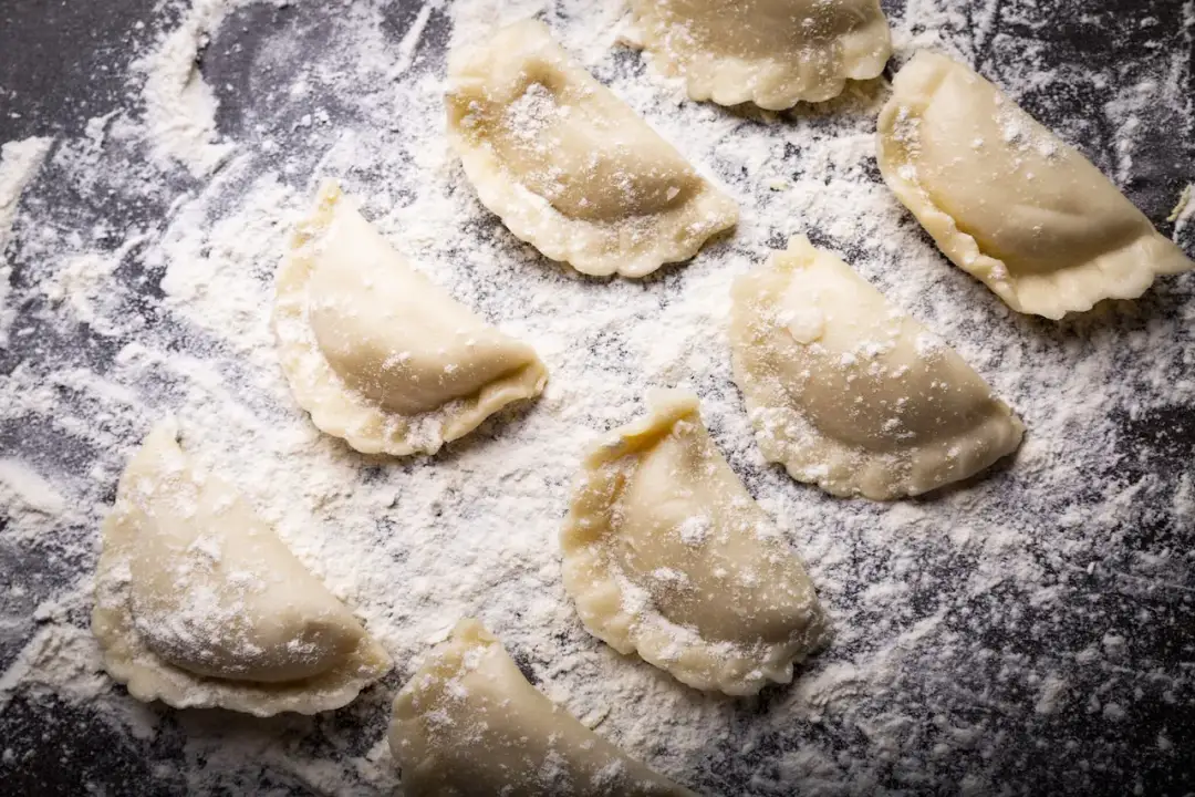 Surowe pierogi leżą na blacie oprószonym mąką. Jaki typ mąki na pierogi? Ta wygląda na pszenną, idealną do domowych wypieków.