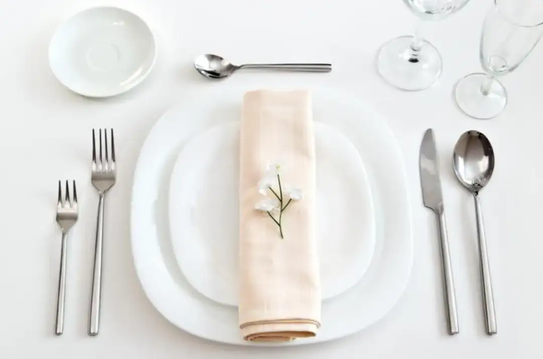 Elegancko nakryty stół: białe talerze, sztućce, serwetka z kwiatkiem i kieliszki.