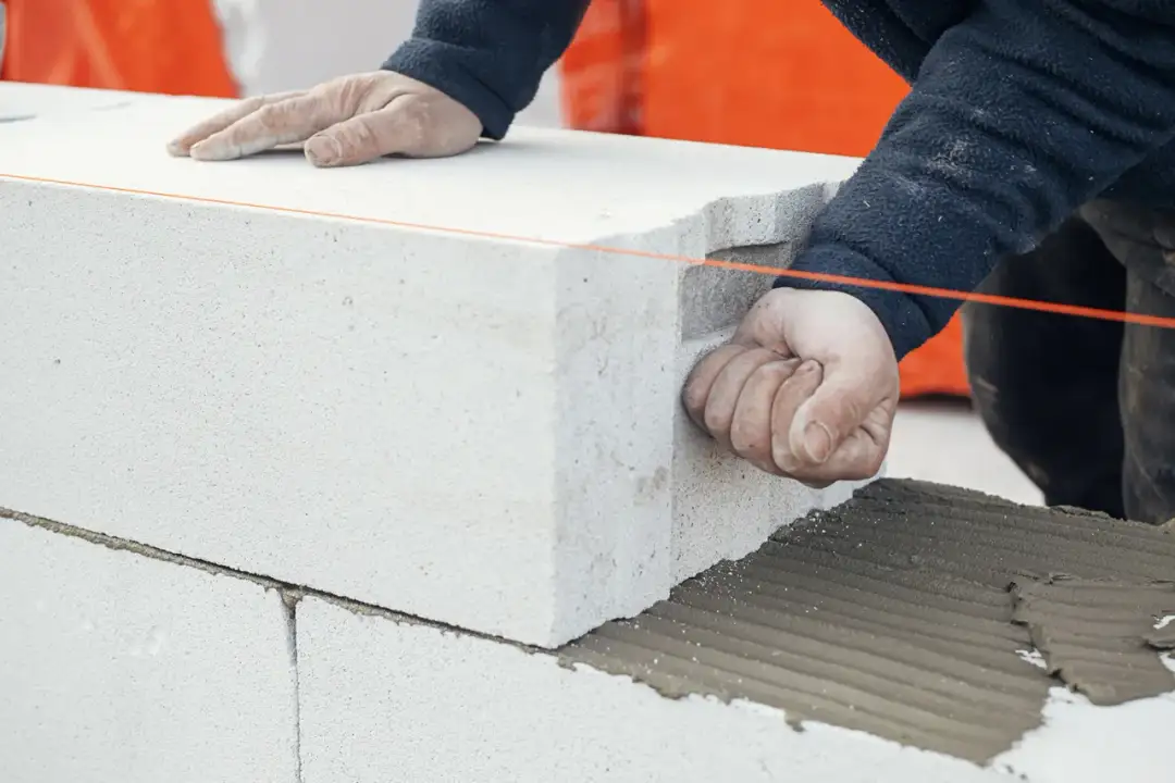 Ręce budowlańca podczas murowania ścianek działowych z bloczków betonowych. Widać zaprawę i sznur murarski.