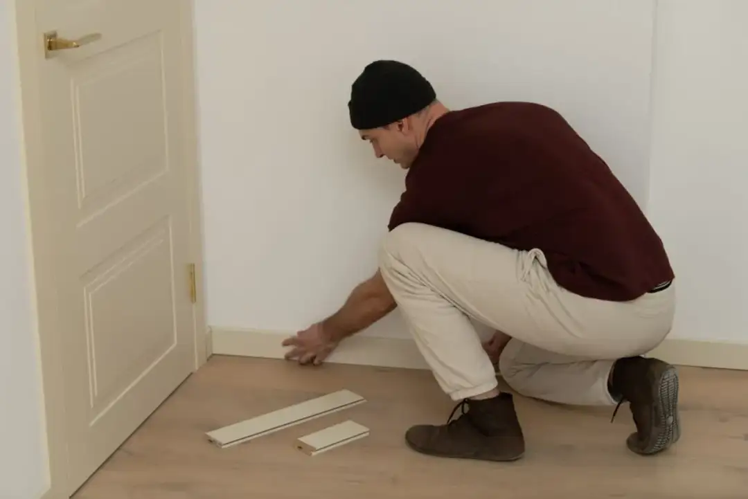 Mężczyzna w czapce i brązowej bluzie montuje listwy przypodłogowe przy drzwiach.
