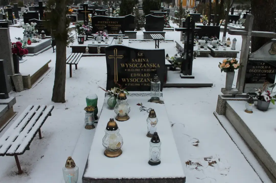 Zimowy cmentarz, grobonet krzyż wlkp. Śnieg przykrywa nagrobki i ławki. Płoną znicze, upamiętniając bliskich.