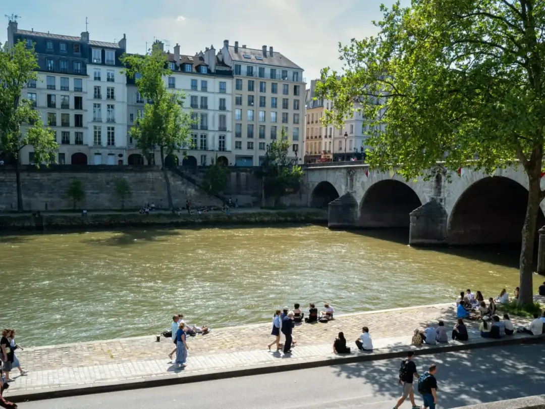 Rzeka, przez którą przepływa Paryż, z mostem i budynkami w tle. Ludzie odpoczywają nad brzegiem.