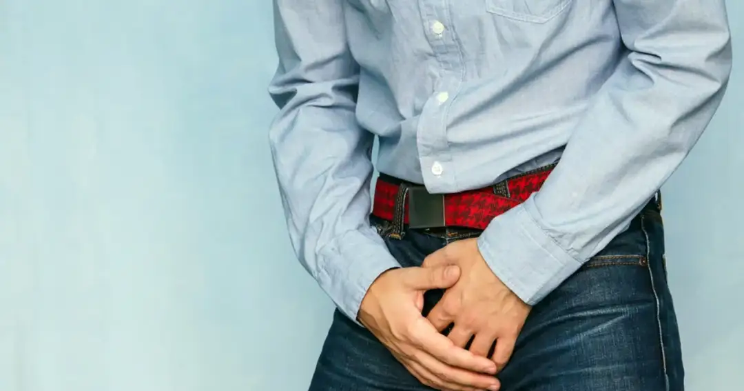 Mężczyzna w niebieskiej koszuli i dżinsach trzyma ręce na kroczu, sugerując problem z wydzieliną z penisa.