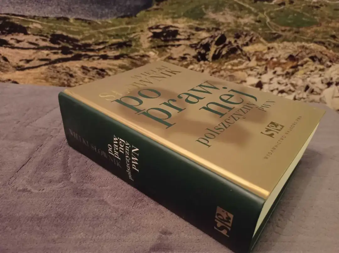 Gruba, złota okładka książki "Wielki słownik poprawnej polszczyzny PWN" leży na szarym materiale.