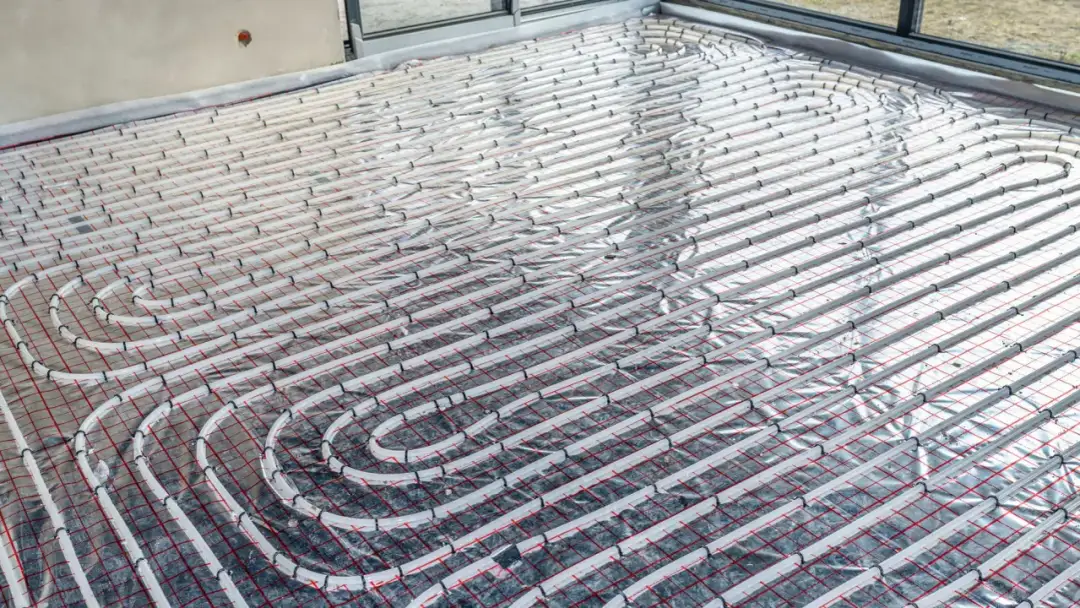 Instalacja ogrzewania podłogowego: białe rurki ułożone na folii izolacyjnej z czerwoną siatką.