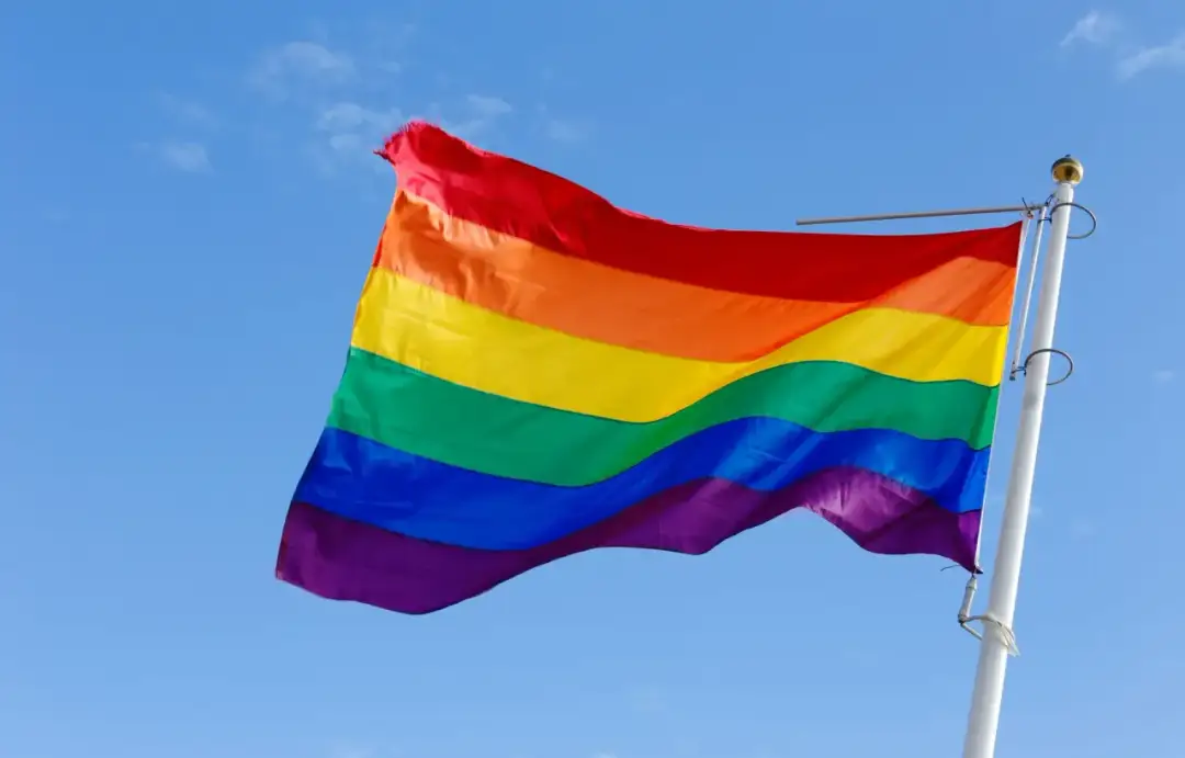 Tęczowa flaga powiewa na maszcie na tle błękitnego nieba. Kolory symbolizują różnorodność i dumę społeczności LGBT.