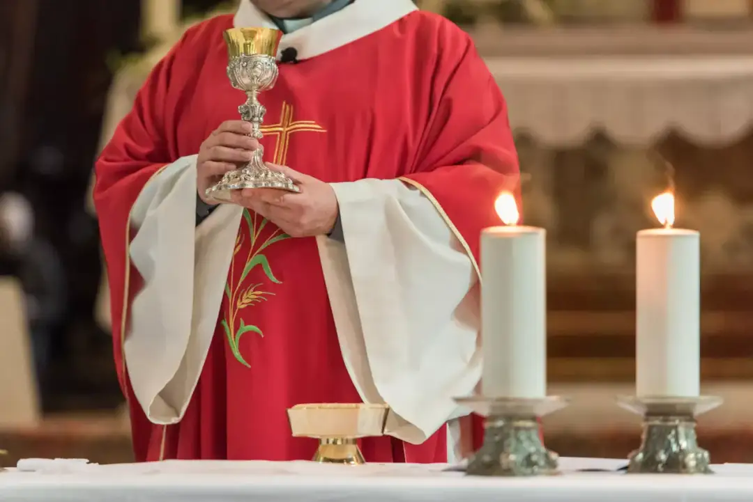 Kapłan w czerwonej szacie z haftem kłosa trzyma kielich podczas modlitwy przed przeistoczeniem. Płoną dwie świece.