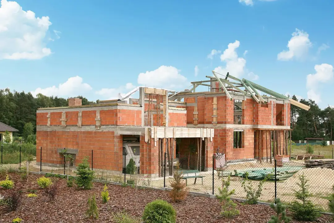 Budowa domu z cegły, widoczne zbrojenie stropu i ścian. W tle las, niebo z chmurami.