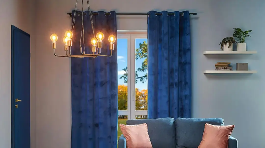 Eleganckie, klasyczne żyrandole do salonu rozświetlają wnętrze. Obok okna z jesiennym widokiem stoi granatowa kanapa.