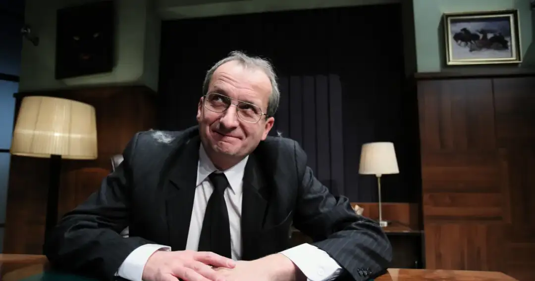 Robert z Kabaretu Moralnego Niepokoju w garniturze, z okularami, zamyślony, siedzi przy biurku.