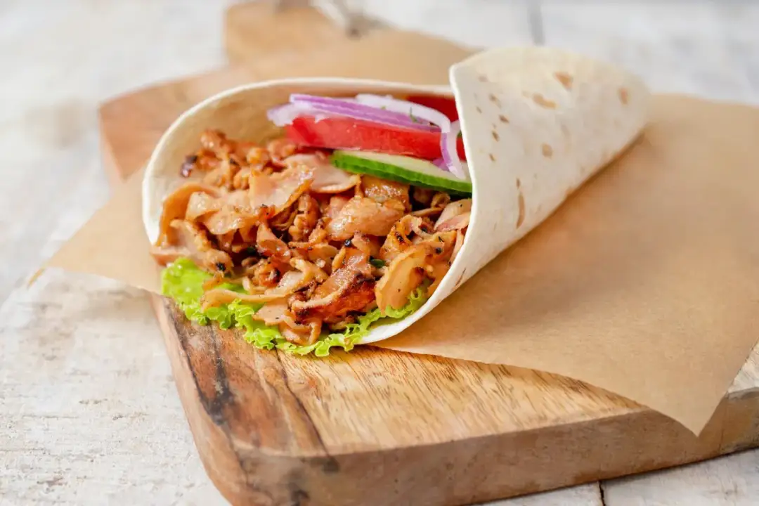 Pyszny kebab w piekarniku, zawinięty w tortillę z soczystym mięsem, sałatą, pomidorem, ogórkiem i czerwoną cebulą, podany na desce.