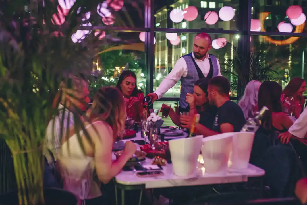 Barka warszawska dzień i noc: kelner nalewa wino gościom przy stole, w tle nocne miasto i dekoracje.