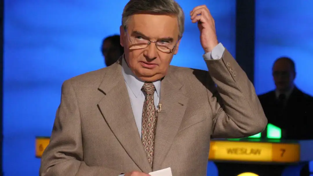 Tadeusz Szmuk, znany z teleturniejów, w garniturze i krawacie, z ręką przy głowie. Czy Tadeusz Szmuk żyje? Tak, jest aktywny.