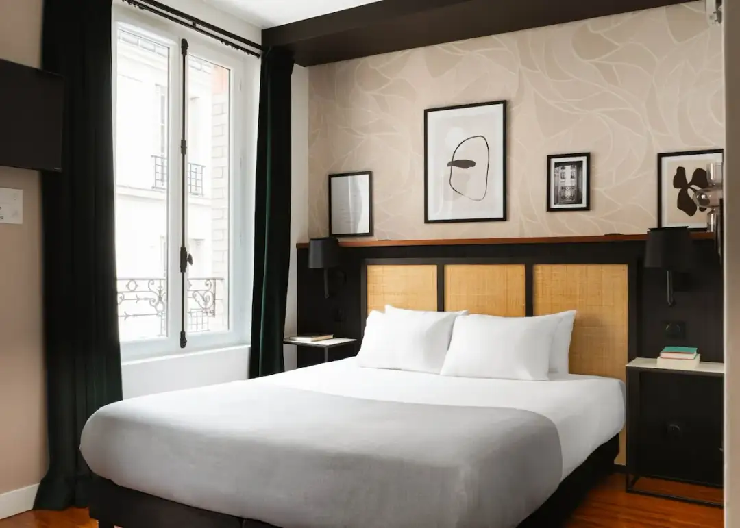 Przytulna sypialnia w 17 dzielnicy Paryża. Łóżko z białymi poduszkami i szarym kocem, okno z widokiem na paryskie kamienice.