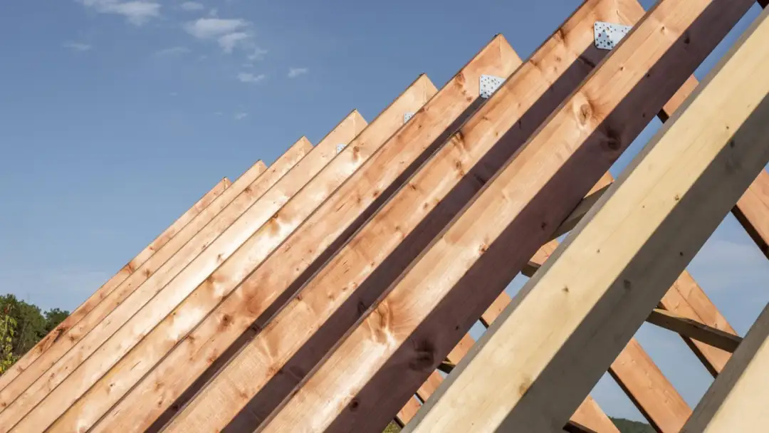 Drewniana konstrukcja dachu pod błękitnym niebem. Widać kolejne krokwie połączone metalowymi kątownikami. Zastanawiasz się, jakie gwoździe do deskowania dachu będą najlepsze?