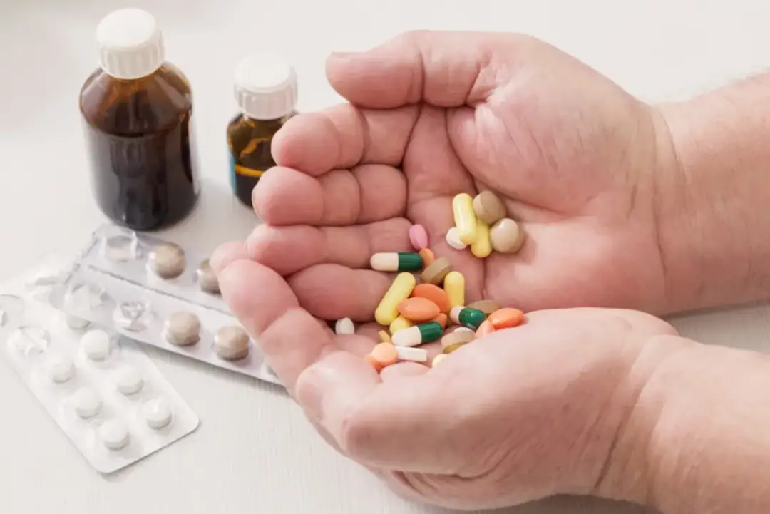 Dłonie trzymające garść kolorowych tabletek i kapsułek, obok buteleczki z lekami i blistry.