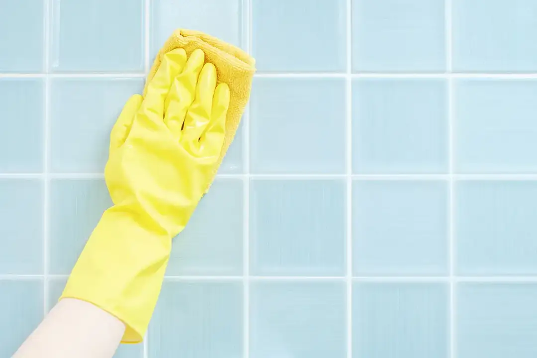 Ręka w żółtej rękawiczce czyści jasnoniebieskie płytki. Dowiedz się, dlaczego profesjonalne sprzątaczki nigdy nie używają octu do mycia fug.