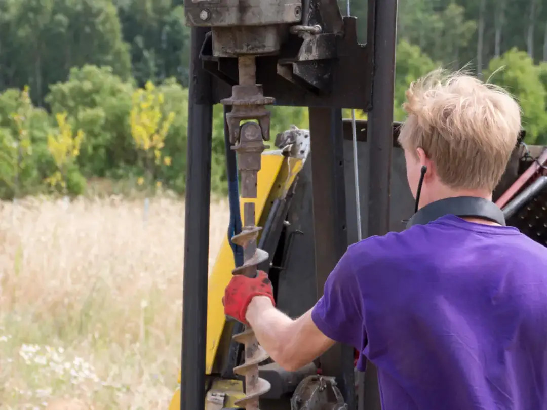 Pracownik w czerwonej rękawicy przygotowuje wiertło do badania gruntu pod budowę domu.